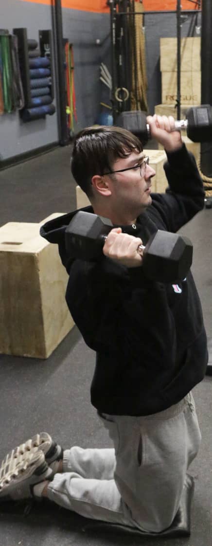 Young member practices kneeling shoulder presses with dumbbells at Apex Performance Gym, NY.