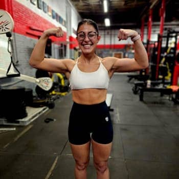 Cheerful female athlete flexing muscles at Apex Performance Gym with gym equipment in the background.