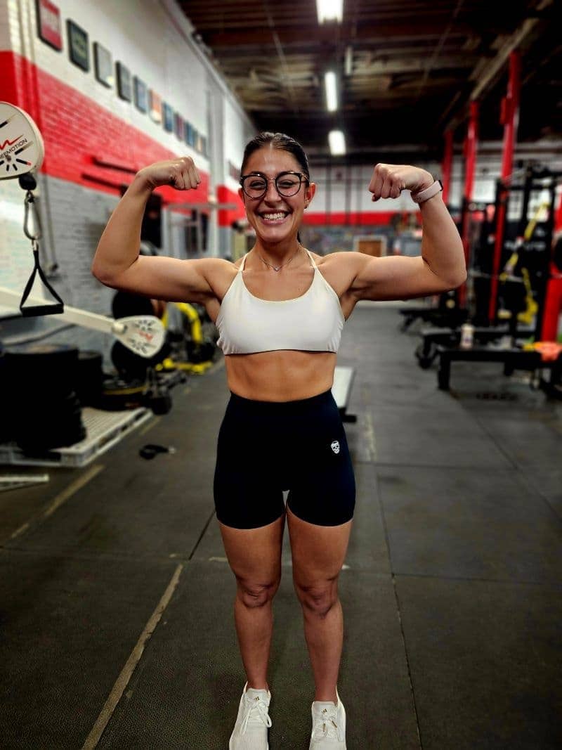 Cheerful female athlete flexing muscles at Apex Performance Gym, Rockland County, with gym equipment in the background.