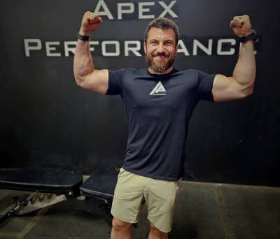 Client flexes muscles proudly at Apex Performance Gym, Rockland County, NY, against branded gym wall.